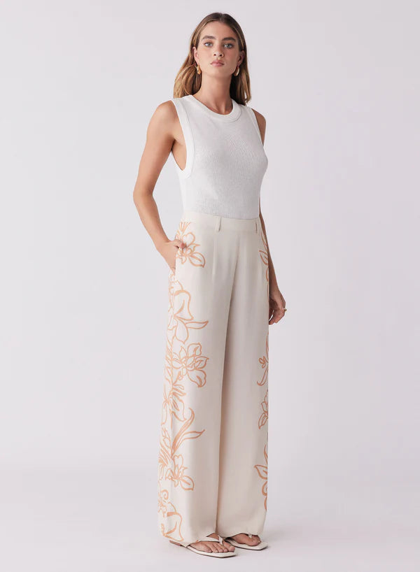 Woman wearing a white sleeveless top and cream pants with floral embroidery on a light gray background
