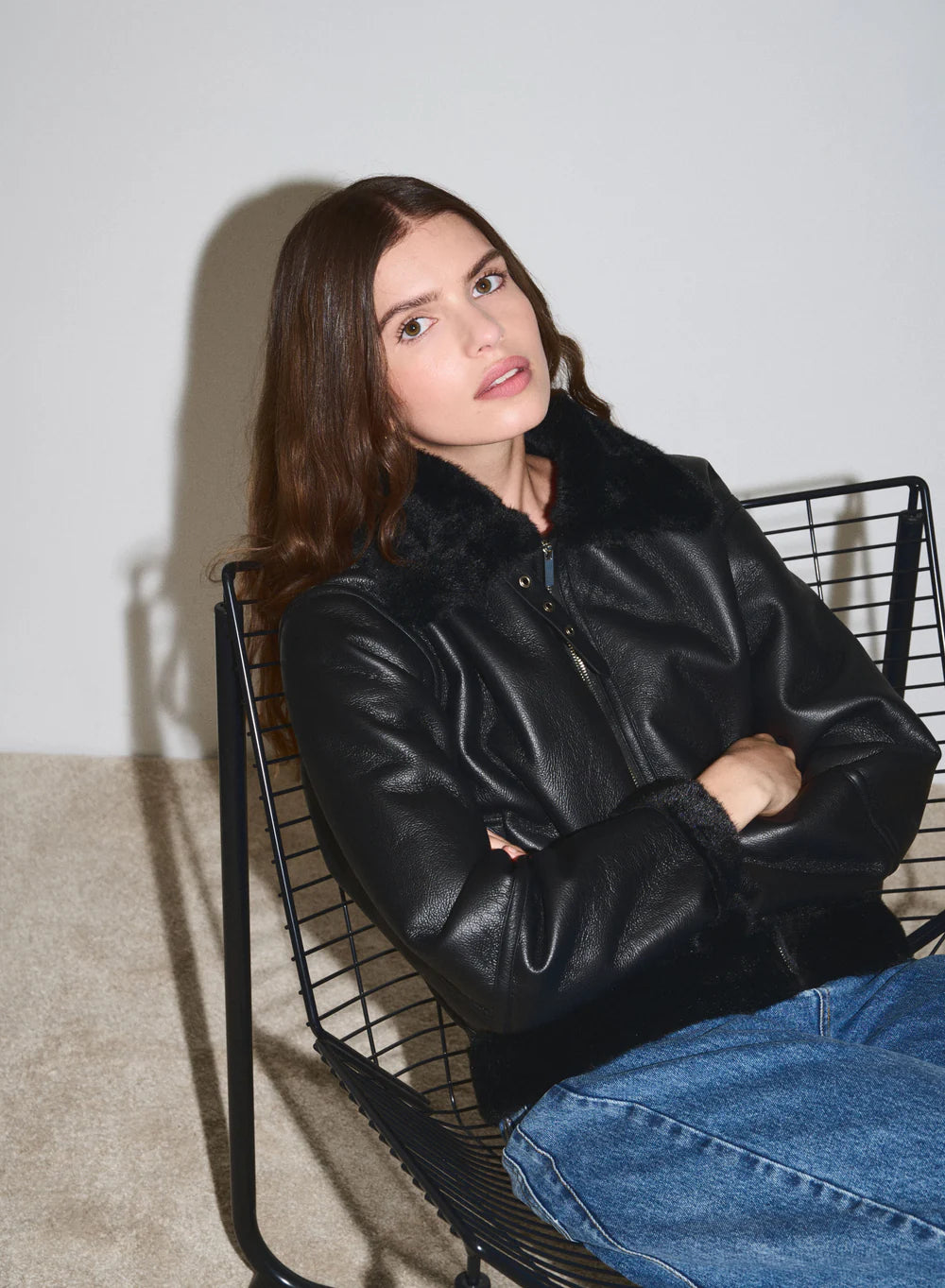 Woman wearing a black leather jacket and blue jeans sitting on a wire chair against a white wall.