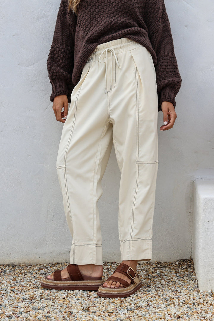 Person wearing ecru btl the label dina pants and brown sandals against a white wall.