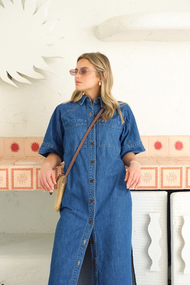 Woman wearing a blue denim dress holding a brown leather bag indoors.