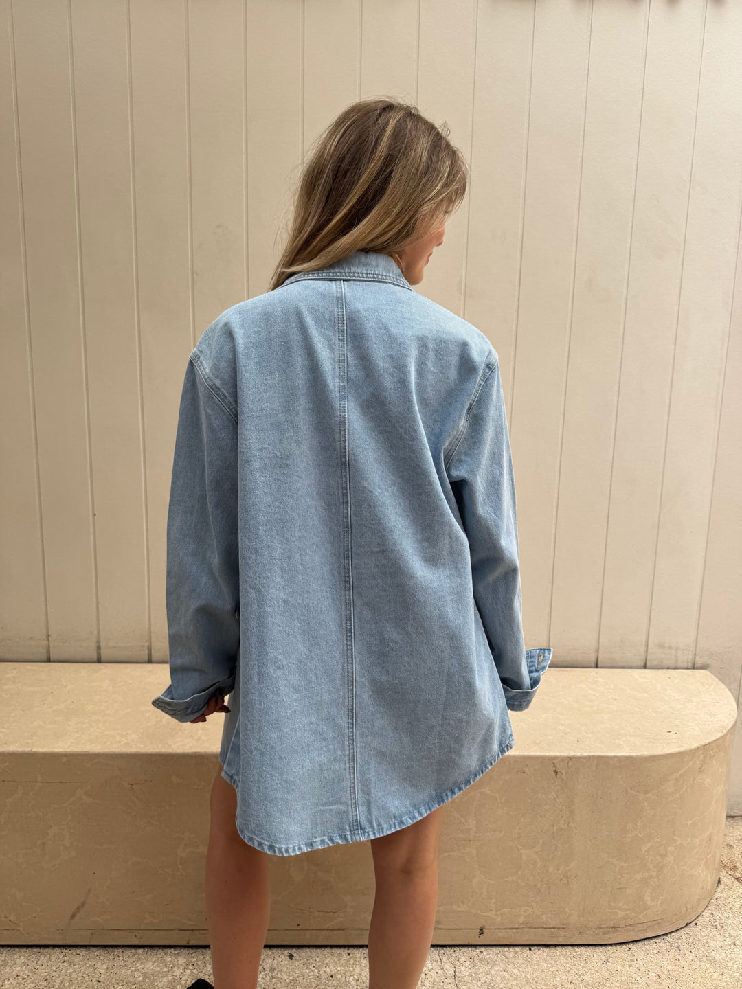 Person wearing a light blue denim shirt standing against a beige wall.