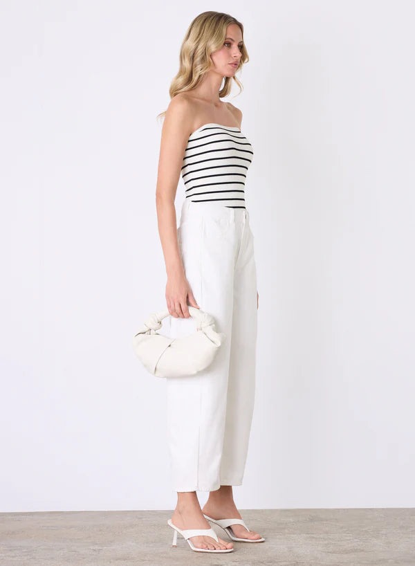 Woman wearing a black and white striped strapless top with white pants, holding a white handbag against a plain background.