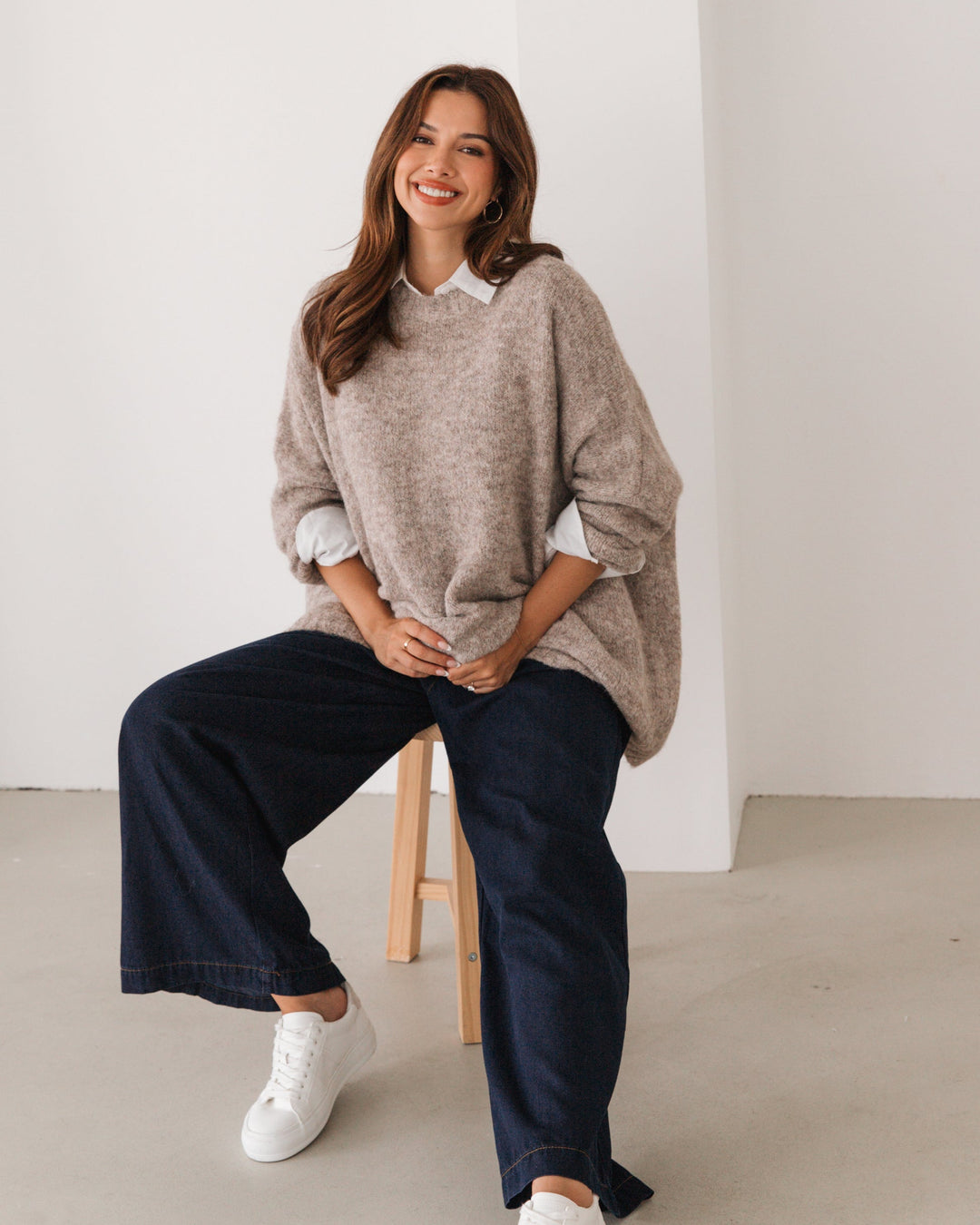 Woman wearing a beige label of love oversize sweater and dark jeans sitting on a stool against a white wall.