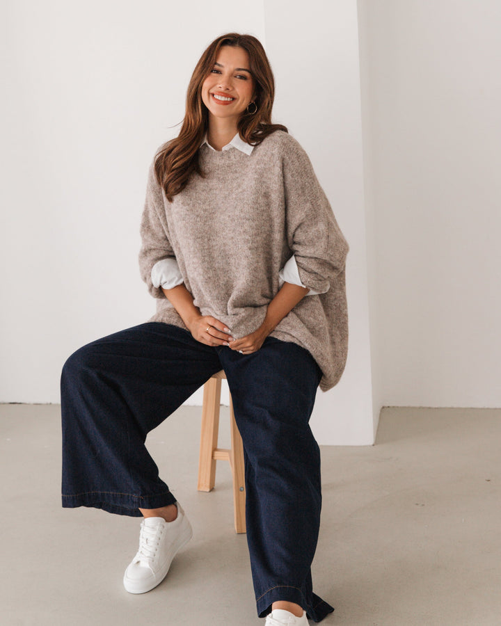 Woman wearing a beige label of love oversize sweater and dark jeans sitting on a stool against a white wall.