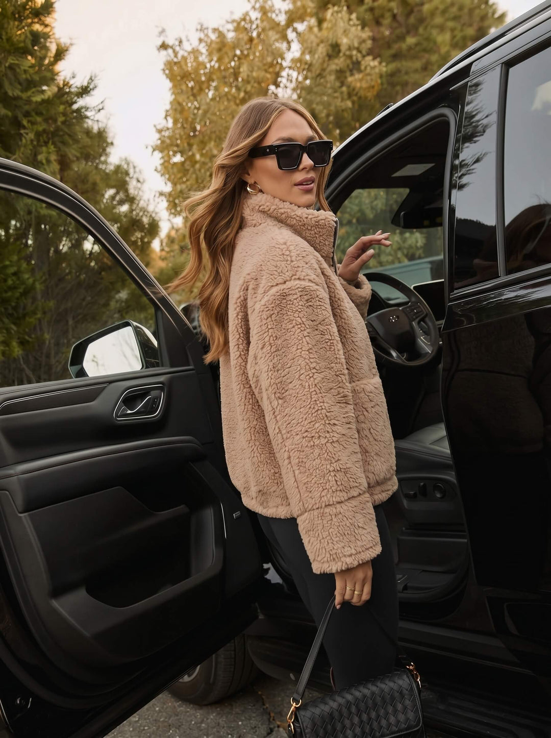 Woman in a beige smaibulun teddy coat getting into a black car with trees in the background