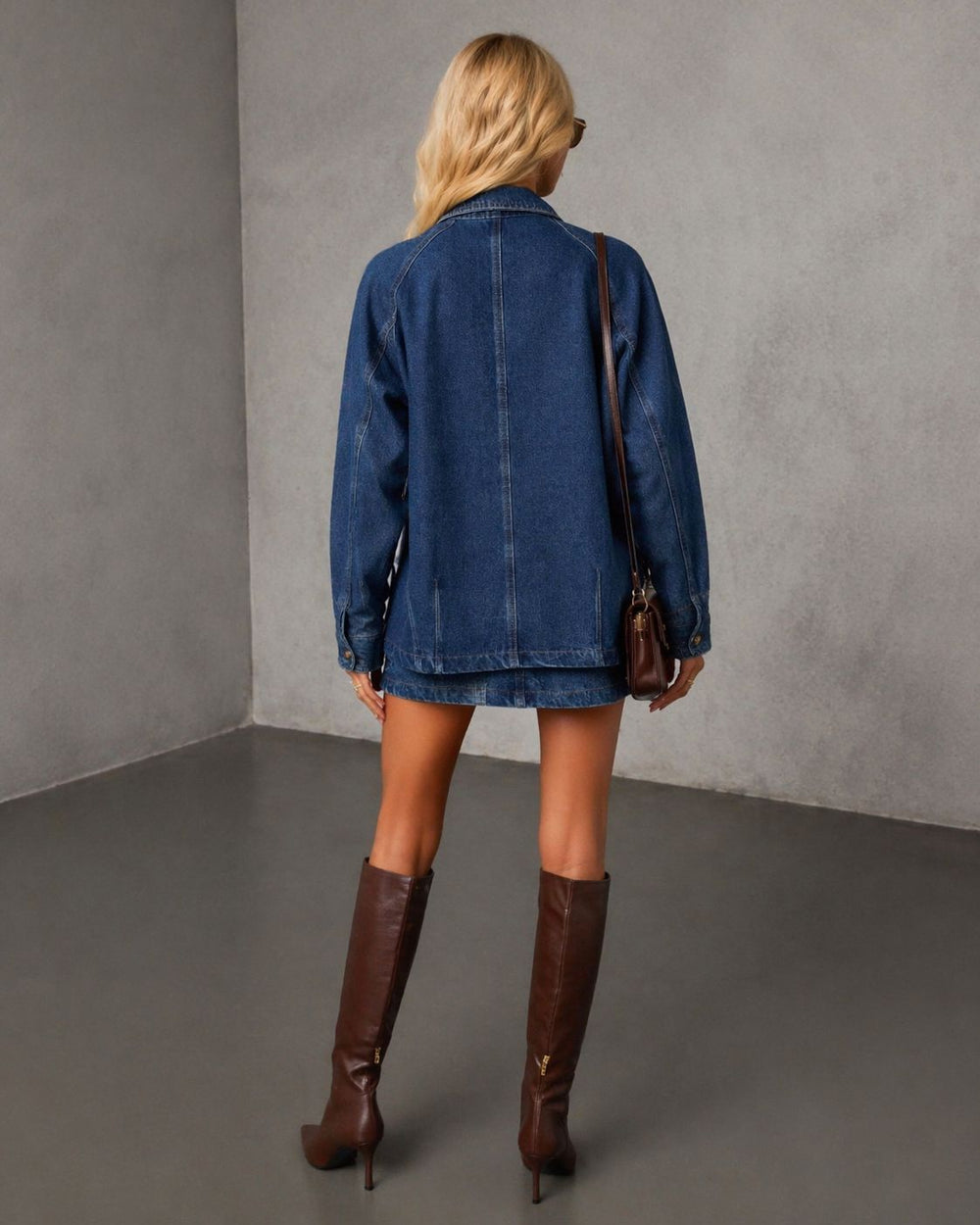 A woman shown from behind wearing the smaibulun denim jacket, brown knee-high leather boots, and carrying a brown shoulder bag in a minimalist gray studio setting.