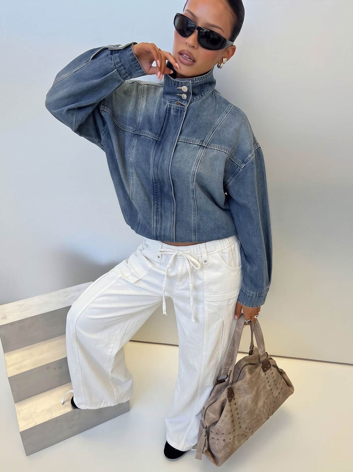 A model wearing a cropped blue smaibulun denim jacket with a high mock neck, white drawstring wide-leg pants, and black sunglasses, holding a taupe textured tote bag while posing on a light gray wooden block against a plain light gray wall.