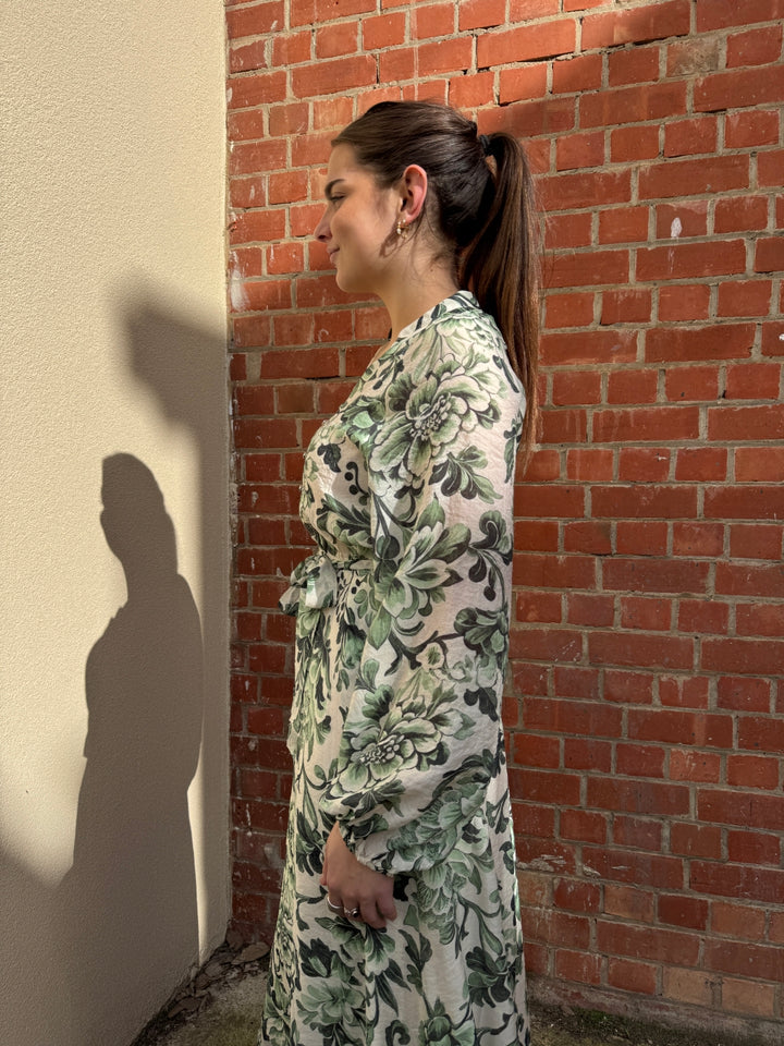 Stella wearing a sunny girl floral dress standing against a brick wall.