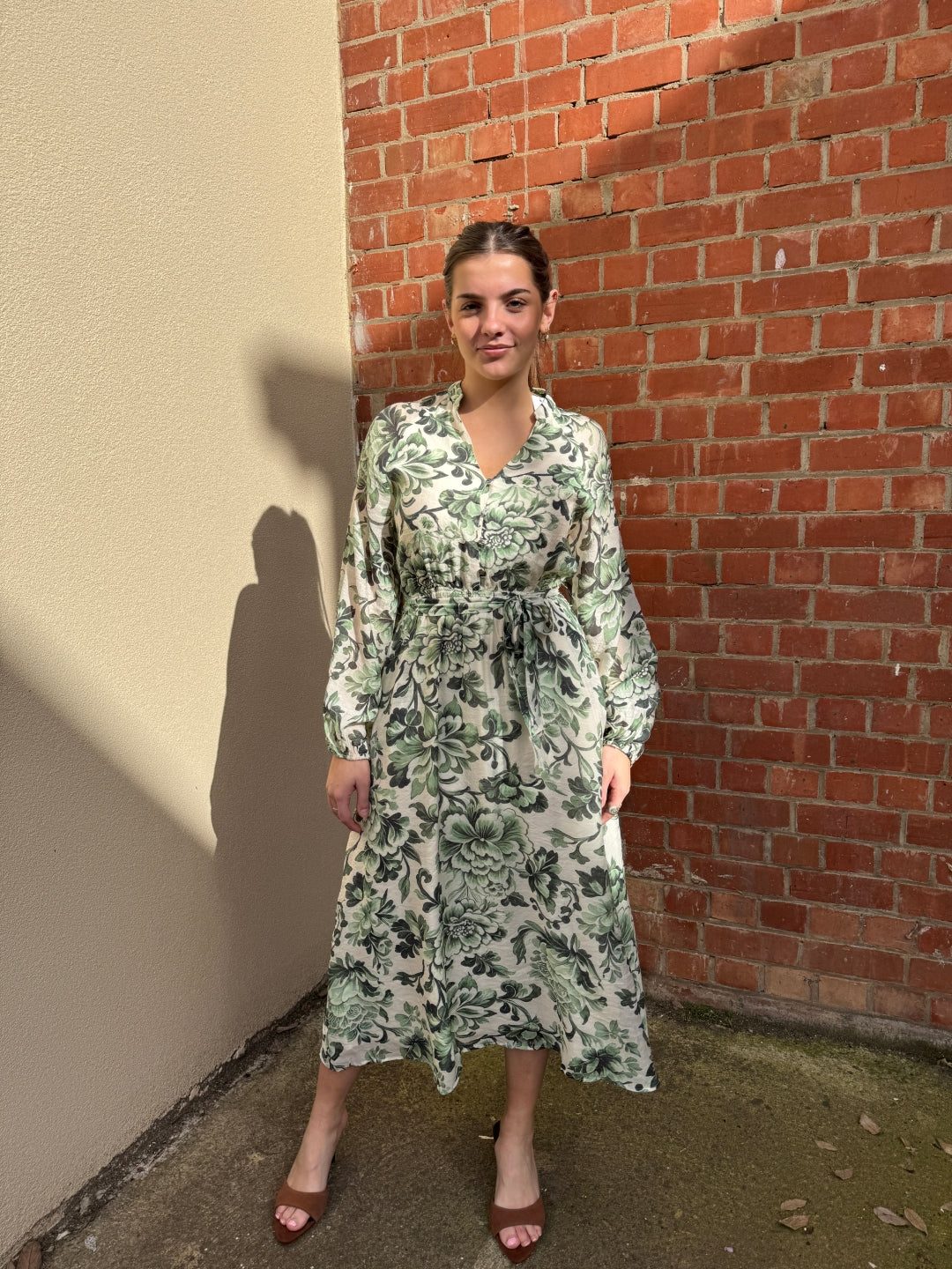 Person wearing a green floral sunny girl dress standing against a brick wall.