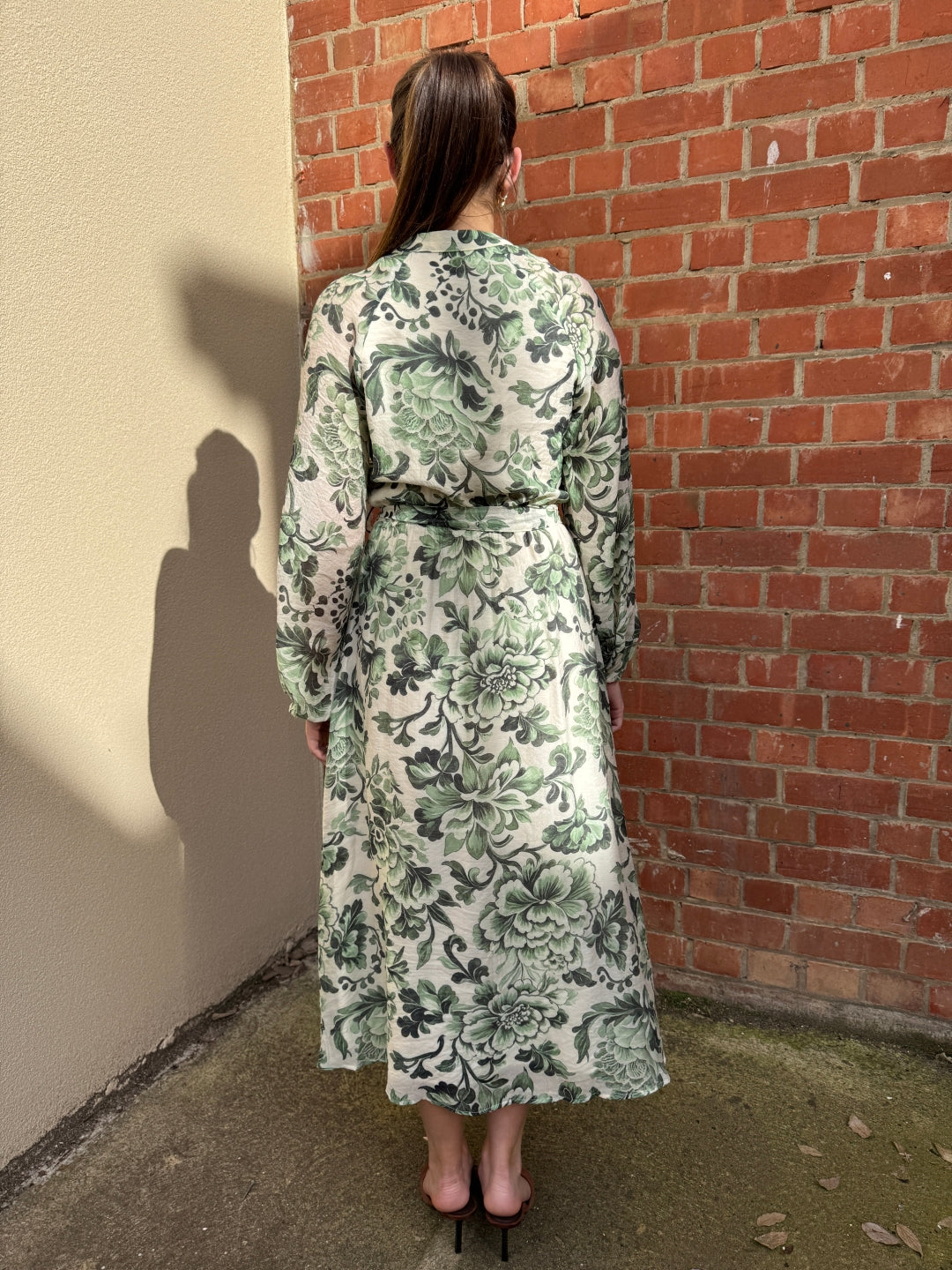 Stella wearing a green floral dress from Sunny Girl standing against a brick wall.