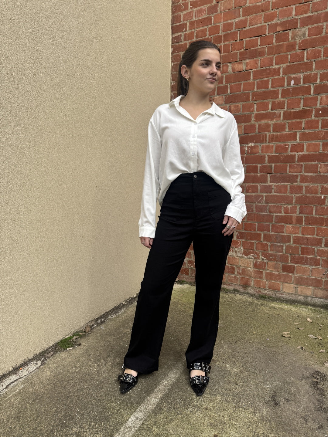 Person wearing a white sunny girl shirt and black pants standing against a brick wall.
