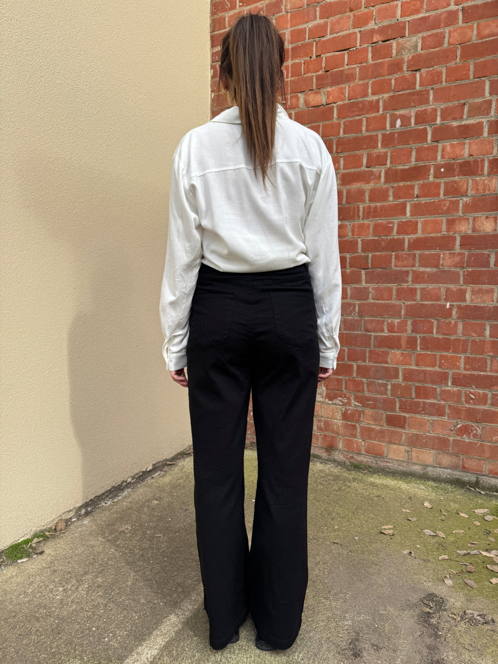 Person wearing a white sunny girl shirt and black pants standing on a sidewalk with a brick wall in the background