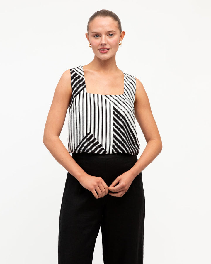A woman modeling a white closet linear luxe sleeveless top with a black and white geometric stripe pattern and a square neckline, paired with black trousers, against a plain white studio background.