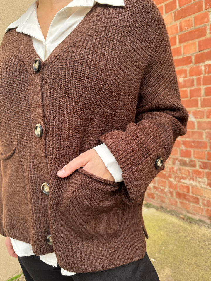 Brown knitted sunny girl cardigan with button details worn by a person, against a brick wall.