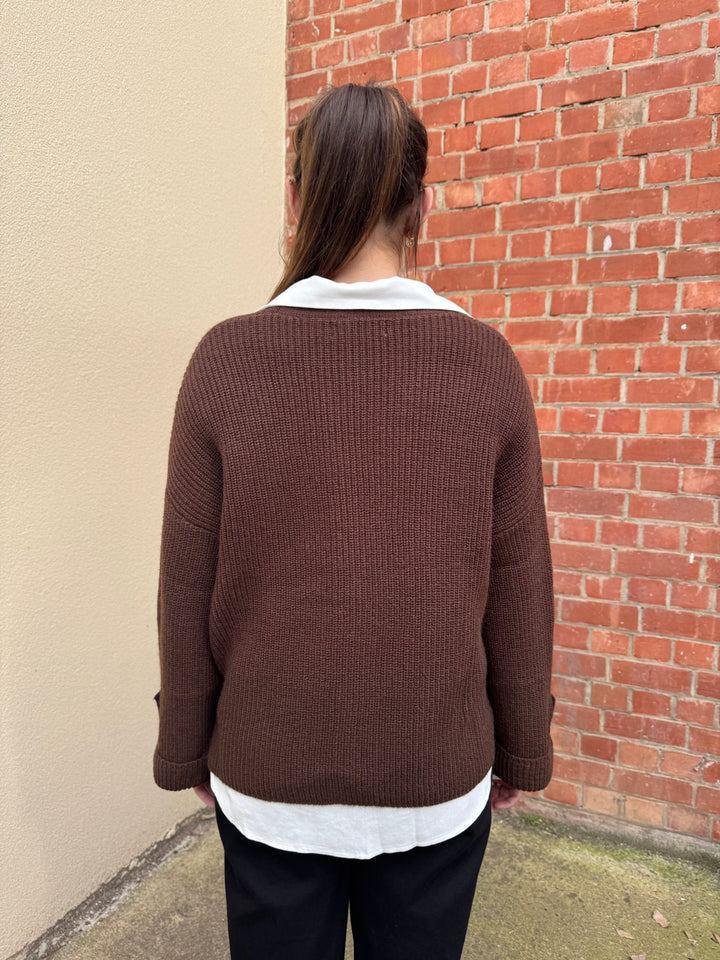 Person wearing a brown sunny girl knit sweater with a white shirt underneath, standing against a brick wall.