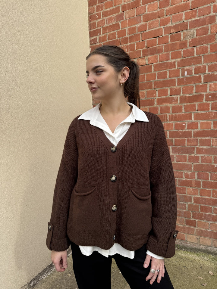 Woman wearing a brown cardigan over a white shirt against a brick wall.
